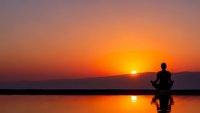 Person meditating in lotus position by calm water during golden sunset with vibrant orange sky reflection creating peaceful zen atmosphere for wellness.