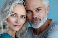 Happy mature couple with silver hair embracing and smiling, showing love and connection in close portrait against blue background.
