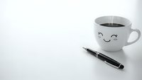 White coffee cup with cute smiling face next to black pen on clean white surface. Minimalist workspace setup for morning routine and productivity concepts.