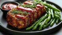 Homemade meatloaf with tomato sauce served on black plate with fresh green beans and rosemary garnish, gourmet comfort food dinner.