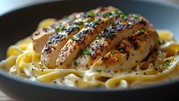 Grilled chicken breast with golden crust served on creamy fettuccine pasta, garnished with fresh herbs and black pepper in dark ceramic plate.