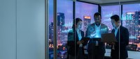 Three diverse business professionals collaborating with tablet in modern office overlooking illuminated city skyline at dusk for corporate teamwork presentations.