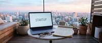 Laptop displaying startup text on desk in high-rise office with city skyline view, potted plants, glasses, and notebook for business planning and entrepreneurship concepts.