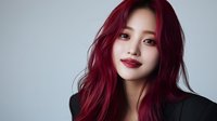 Young Asian woman with vibrant burgundy red hair and red lipstick smiling confidently against gray background wearing black blazer, professional beauty portrait.
