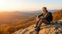 Male hiker with brown hair and light skin tone sitting on rocky mountain summit at golden hour, overlooking vast forested valley and distant peaks during sunset or sunrise adventure.