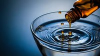 Golden oil pouring from amber bottle into clear glass creating ripples and droplets against blue gradient background for wellness and beauty concepts.