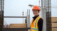 Smiling female construction worker in orange hard hat and safety vest at building site with concrete structures and rebar in background.
