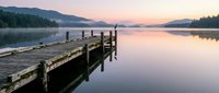 Wooden dock extending into calm lake at sunrise with misty mountains and forested shoreline reflected in still water. Serene landscape for travel, nature, and relaxation themes.