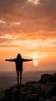 Silhouette of woman with arms outstretched standing on mountain peak during golden sunset with dramatic orange sky and rolling hills in background.