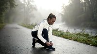 Young adult male, South Asian ethnicity, wearing casual athletic attire, tying shoelaces on misty forest road near water. Suitable for fitness, wellness, and outdoor lifestyle content.