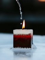 Water drops falling onto burning red candle in snow creating magical winter atmosphere with flame reflection and frozen crystals around base.