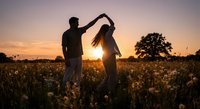 Romantic couple dancing in golden sunset field with wildflowers, silhouetted against warm evening sky for wedding, love and relationship concepts.