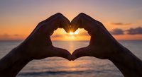 Hands forming heart shape silhouette against golden sunset over ocean horizon creating romantic scene for love and relationship concepts.