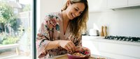 Young blonde woman in floral robe preparing healthy breakfast bowl with granola and berries in bright modern kitchen, morning wellness routine lifestyle.