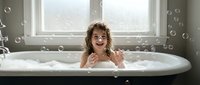 Happy young Caucasian girl with curly hair enjoying bubble bath in white bathtub by window, smiling and playing with soap bubbles in bright bathroom.