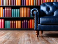 Navy blue leather armchair with tufted back and nailhead trim against colorful library bookshelf background on wooden floor, creating cozy reading atmosphere.