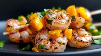 Grilled shrimp skewers with fresh mango chunks and herbs on dark plate, close up view of seafood appetizer garnished with chopped parsley and spices.