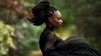 Young woman with dark skin tone and braided hair styled in an updo, wearing black flowing fabric, photographed in profile against a blurred green natural background for lifestyle and fashion content.