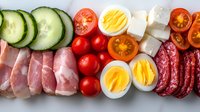 Fresh ingredients arranged in rows including sliced cucumber, ham, cherry tomatoes, boiled eggs, feta cheese and salami on white background, top view food photography.