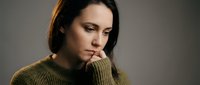Thoughtful young woman in olive green sweater looking down with contemplative expression against neutral background for lifestyle and emotional wellness content.