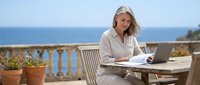 Middle-aged woman with long gray hair, Caucasian ethnicity, working remotely on laptop at outdoor terrace table overlooking Mediterranean sea. Freelancer or entrepreneur using computer for business tasks.