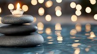 Zen stones pyramid with burning candle on top against blurred bokeh lights reflecting in water surface, creating peaceful spa atmosphere.