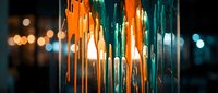 Modern pendant lamp with orange and teal glass strands cascading downward, illuminated against dark blurred background with bokeh lights, suitable for interior design and lighting projects.