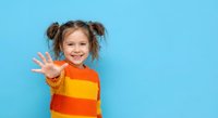 Happy young girl with pigtails waving hand in colorful striped sweater on blue background. Cheerful child greeting gesture for education and childhood concepts.