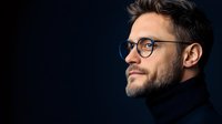Profile view of confident Caucasian man with beard wearing glasses and turtleneck sweater against dark background, dramatic studio lighting creates professional atmosphere.