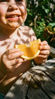 Young child with medium skin tone enjoying fresh orange slice outdoors, smiling brightly while holding citrus fruit in hands. Summer snack and healthy eating concept.