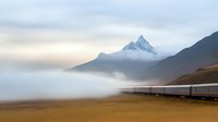 Scenic railway journey through misty mountain landscape with snow-capped peak rising above low-lying clouds, train cars stretching into foggy distance at sunrise.