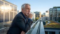 Mature male professional with gray hair and light complexion, wearing dark blazer, standing on urban rooftop terrace overlooking city skyline. Thoughtful pose with hand near face, contemplating business strategy or future plans.