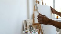 Male artist with dark skin tone holding blank canvas on easel in bright studio space with painting supplies and natural light for creative work and art education.