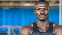 Young athletic African American man in black tank top at gym, confident expression against blue background, muscular shoulders and strong facial features.