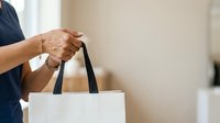 Man holding white shopping bag with black handles, closeup of hands and arms in casual setting for retail and commerce concepts.