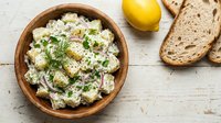 Creamy potato salad with fresh dill and red onion in wooden bowl, served with lemon and whole grain bread slices on white table.