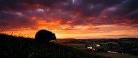 Dramatic sunset landscape with vibrant orange and purple clouds over rolling hills and countryside valley with scattered lights below.