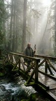 Middle-aged man with long hair and beard, wearing olive jacket and carrying bow, standing on wooden bridge over misty forest stream surrounded by tall evergreen trees.