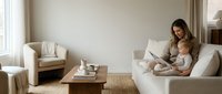 Young mother with toddler daughter using laptop computer while sitting together on white sofa in modern minimalist living room interior for remote work.