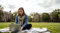 Young Caucasian female college student studying outdoors on campus lawn with laptop and textbooks during golden hour for academic success.