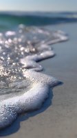 Gentle ocean wave with white foam washing onto sandy beach at sunrise, creating sparkling bokeh effects and soft reflections in morning light.
