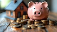 Pink ceramic piggy bank with stacked coins and miniature house on wooden surface, representing savings, investment and real estate financial planning.