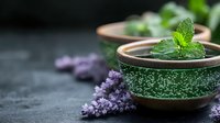 Fresh mint leaves in ceramic bowls with purple lilac flowers on dark stone background, aromatic herbs for tea and natural medicine preparation.