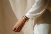 Female hand with multiple gold bracelets wearing elegant white dress against neutral beige background, showcasing luxury jewelry accessories.