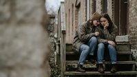 Young couple in casual winter clothing sitting together on stone steps of brick building sharing intimate moment outdoors in urban setting.