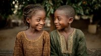 Two happy African children smiling and laughing together outdoors in natural sunlight wearing traditional colorful clothing in warm earth tones.