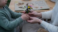 Adult daughter giving small wildflower bouquet to elderly mother on Mother's Day, hands exchanging flowers with handmade card on wooden table background.
