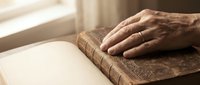 Elderly hand resting on vintage leather-bound book by window with soft natural light creating peaceful reading moment for literature and wisdom concepts.