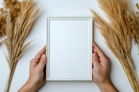 Hands holding white frame mockup with dried pampas grass decoration on neutral background. Minimalist design layout for artwork presentation.