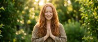 Young redhead woman with closed eyes in prayer pose outdoors during golden hour sunset, peaceful meditation moment in natural green forest setting.